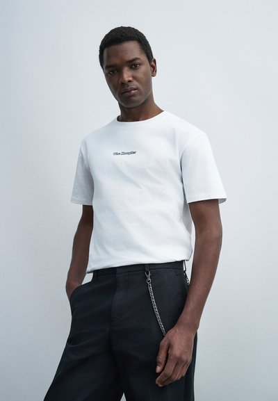 Νεαρός άνδρας με λευκό T-shirt με το λογότυπο "The Kooples" και μαύρο παντελόνι με ασημένια αλυσίδα, σε μονόχρωμο ανοιχτό γκρι φόντο.