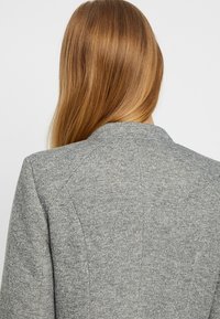 Harmaa bleiseri, jossa on teksturoitu kangas, jämäkät olkapäät ja hapsureunainen kaulus. Ominaisuuksiin kuuluu muotoiltu takasuunnittelu ja minimaalinen ompelutyyli.