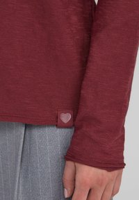 Weinrotes Langarmshirt aus strukturiertem Stoff mit einem kleinen herzförmigen Etikett am Saum und körpernahen Bündchen. Kombiniert mit grauen Nadelstreifen-Hosen.