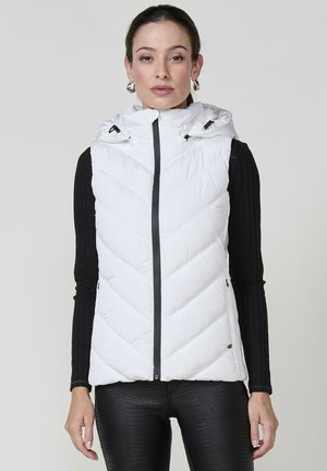 Chaleco blanco acolchado con capucha, con costura en forma de espiga, cierre frontal con cremallera y cordones ajustables. Camisa de manga larga negra acanalada debajo.