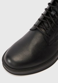 Pantof cu piele neagră, având o suprafață texturată, vârf rotund și un design cu șireturi, cu cusături contrastante și o talpă din cauciuc rezistentă.