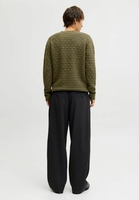 Person standing facing away, wearing an olive green textured sweater and loose black pants against a plain white background.