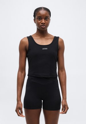 Γυναίκα που φοράει μαύρο κοντό τοπ με το λογότυπο "GUESS" στο μπροστινό μέρος και μαύρα, εφαρμοστά σορτς με ψηλή μέση, στέκεται μπροστά σε λευκό φόντο.