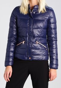 Mörkblå puffjacka med ett kviltat mönster, två fickor med dragkedja framtill och detaljer i roséguld. Slät, glansig textur.