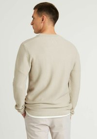 Maglione beige texture con scollatura rotonda e maniche lunghe, con polsini e orlo a costine. Indossato sopra pantaloni chiari.