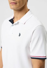 Balta polo marškinėliai su juodos ir raudonos spalvos dryžuota apykakle, su mažu siuvinėtų logotipu ant krūtinės. Pagaminti iš tekstūruotos medžiagos.
