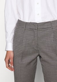 Personne portant un pantalon sur mesure à motif pied-de-poule et une chemise blanche à manches longues boutonnée, main détendue sur le côté.
