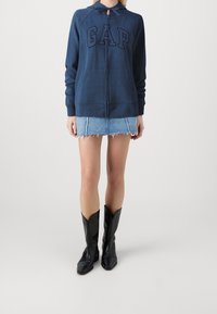 Sudadera con cremallera en color navy con el logo "GAP", combinada con una falda corta de mezclilla clara con bordes deshilachados y botas de cowboy negras.