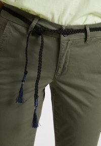 Pantalon vert olive avec une ceinture noire tressée ornée d'accents bleu marine et de pompons, fermeture à boutons à l'avant et détail de poche.
