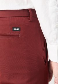 Bordowe spodnie o gładkiej fakturze, z tylną kieszenią i szlufkami na pasek. Logo "BOSS" znajduje się na czarnej naszywce.