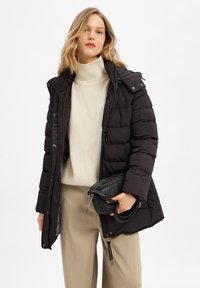 Czarna kurtka puchowa z zamkiem z przodu, wysokim kołnierzem i odpinanym kapturem. Nosi na sobie kremowy golf i beżowe spodnie, trzymając w ręku małą czarną torbę.