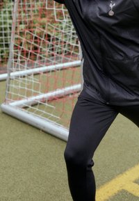 Svart sportjacka med logotyp, kombinerad med svarta leggings. Bakgrunden har en målstolpe och konstgräs.