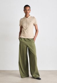 Beige polo skjorte med tre knapper, kombinert med løse, teksturerte olivengrønne bukser, og svarte spisse sko. Enkel design, nøytrale farger.