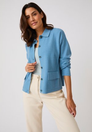 Mujer que lleva un cárdigan azul claro abotonado sobre una camiseta azul pálido y pantalones beige, de pie frente a un fondo liso y claro.