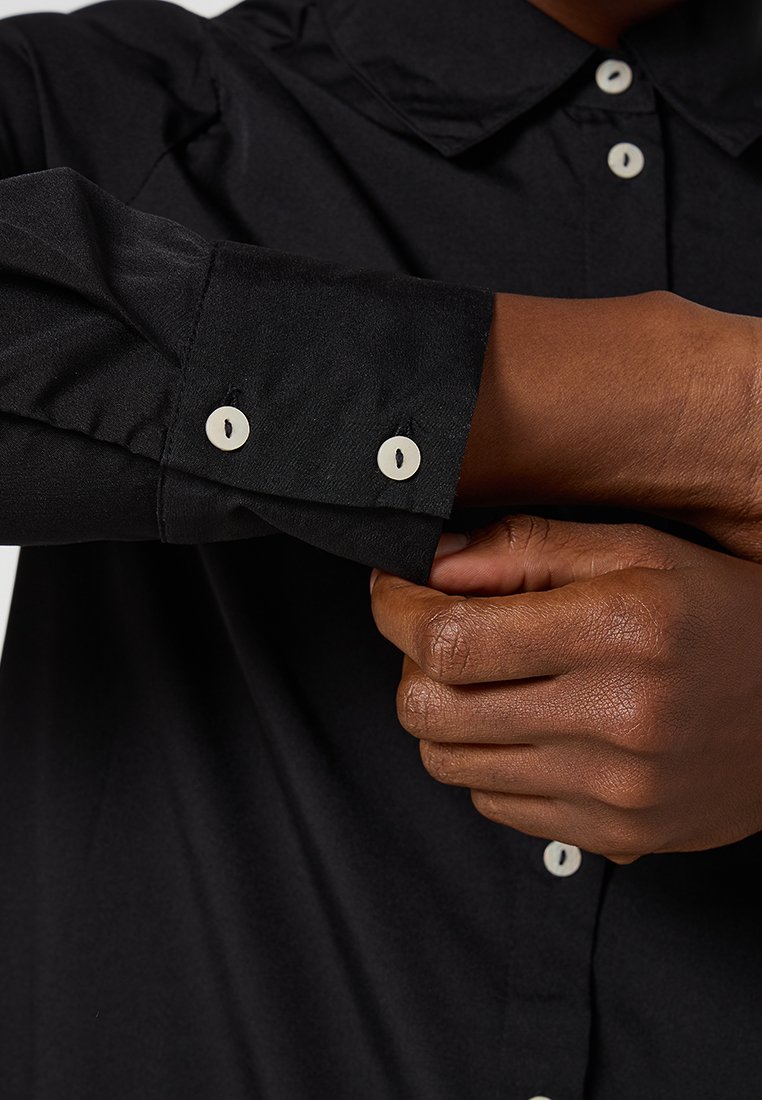 Zwarte button-up overhemd met een puntige kraag en witte knopen. De stof heeft een gladde textuur, met een close-up van een manchetdetail.