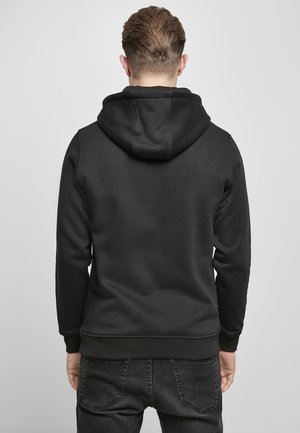 Homme aux cheveux bruns courts portant un sweat à capuche noir et un jean foncé, de dos sur un fond clair uni.
