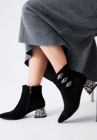Botines de ante negros hasta el tobillo con punta afilada, que cuentan con un cierre de cremallera plateado y tres detalles ovalados. Los tacones de bloque presentan un diseño estampado.