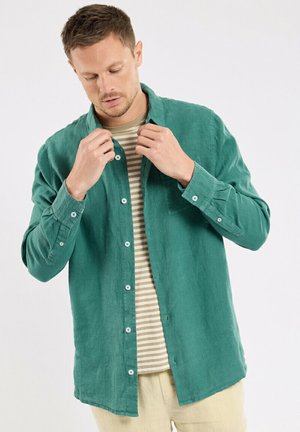 Homme boutonnant une chemise à manches longues couleur sarcelle par-dessus un t-shirt rayé beige et blanc, portant un pantalon beige clair, sur fond blanc.