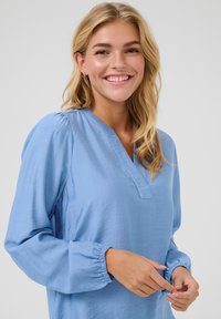 Blusa azzurro chiaro realizzata in tessuto testurizzato, con scollo a V, maniche arricciate e polsini elasticizzati. Design semplice senza motivi.