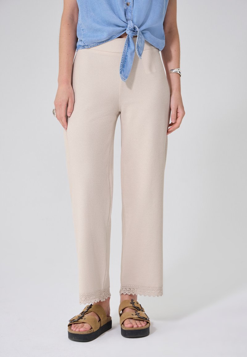 Femme portant un pantalon en maille beige avec une bordure en dentelle, des sandales compensées marron clair et une chemise sans manches bleue nouée, debout sur un fond clair.