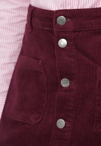 Burgundi bordázott nadrág, függőleges bordázott textúrával, két elülső zsebbel és ezüst színű patentgombokkal.
