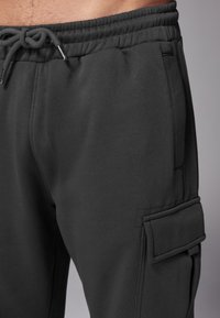Mörkgrå sweatpants har en dragsnodd i midjan, sidofickor och en cargoficka på vänster ben med klaffdetalj. Slät textur.