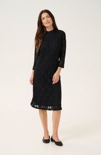Robe en dentelle noire avec des manches trois-quarts, motif floral et un ourlet accentué par de la dentelle transparente. Associée à des chaussures plates noires.