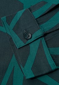 Colletto di camicia verde con motivo su sfondo scuro. Presenta un bottone nero al colletto e bold geometrico in turchese.