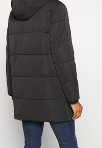 Osoba v černé péřové bundě s kapucí a bočním zipem, v kombinaci s modrými džíny, stojící na jednobarevném pozadí.