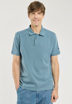 Homme souriant portant un polo bleu et un jean bleu, les mains dans les poches devant un fond uni.