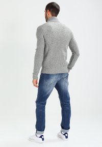 Man som bär en grå stickad polotröja, blå uppvikta jeans och vita sneakers, står vänd bort från kameran mot en neutral bakgrund.