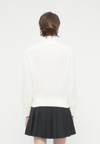 Λευκό π�λεκτό πουλόβερ με ψηλό λαιμό και ραμμένο στρίφωμα, συνδυασμένο με πλισέ φούστα σε σκούρο γκρι χρώμα, τοποθετημένο σε απλό φόντο.