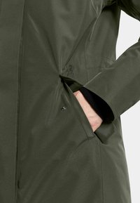 Olijfgroen waterdichte jas met een gladde textuur, zijzak met drukknoopsluiting en een geborduurd merkimago nabij de zak.