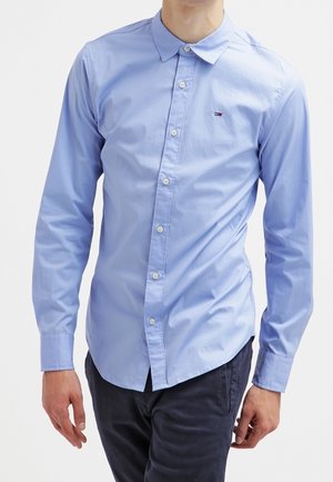 Homme portant une chemise cintrée bleu clair à manches longues avec un petit logo sur la poitrine et un pantalon foncé.