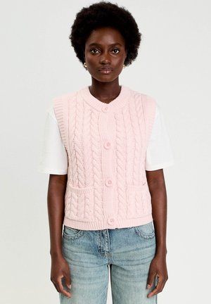 Giovane donna con capelli corti e ricci che indossa un gilet rosa a trecce con bottoni sopra una camicia bianca a maniche corte e jeans blu, su uno sfondo neutro.