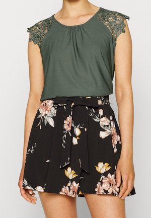 Top verde scuro con dettagli in pizzo sulle spalle, abbinato a pantaloni corti neri con fiori e cintura annodata. Tessuto morbido e vestibilità rilassata.