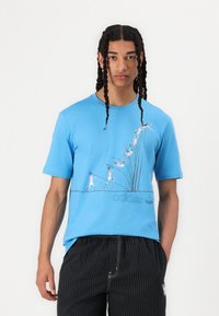 Kék, rövid ujjú póló mozgásban lévő alakokkal és kontrasztos színű "adidas" logóval díszítve. Pamut anyag, laza szabású.