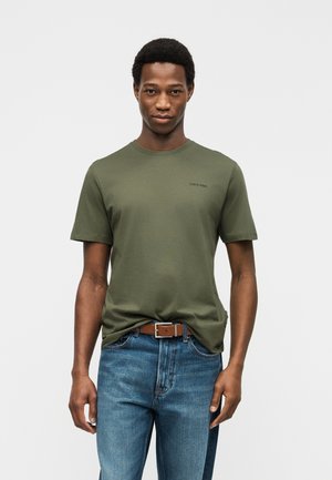 Hombre vestido con una camiseta verde oliva de Calvin Klein, jeans azules y un cinturón marrón, de pie mirando al frente contra un fondo liso.