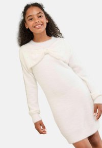 Weißes gestricktes Kleid mit langen Ärmeln, weichem Material und einer Schleifen-Detail am Ausschnitt, akzentuiert mit kleinen, funkelnden Verzierungen.