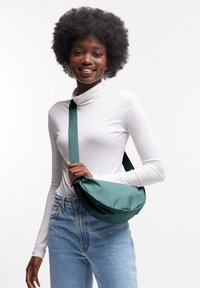 Borsa a tracolla verde in satin con forma curvo e tracolla regolabile, tenuta da una persona in un dolcevita bianco e jeans blu.