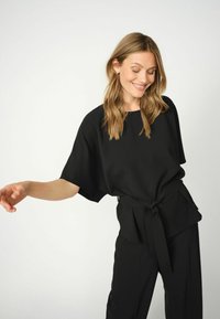 Top nero, dal taglio morbido e maniche corte, con vita annodata. Tessuto liscio e materiale leggero, abbinato a pantaloni coordinati.