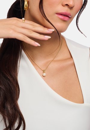 Nahaufnahme einer Frau mit goldenen Creolen, einer goldenen Halskette mit einem Perlenanhänger und einem goldenen Ring mit einer Perle.