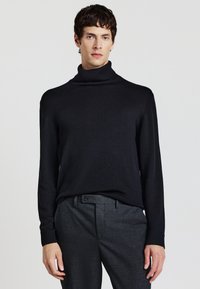 Maglione nero a collo alto con polsini e colletto a costine, abbinato a pantaloni scuri dalla texture particolare. Il materiale sembra morbido e lavorato a maglia.