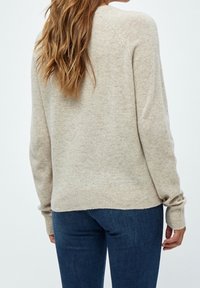 Kvinna med långt vågigt hår, klädd i en beige stickad tröja och mörkblå jeans, visad bakifrån mot en enfärgad bakgrund.