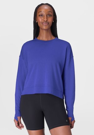 Femme aux cheveux tressés portant un sweat-shirt bleu à manches longues et un short de cycliste noir, debout devant un fond clair uni.