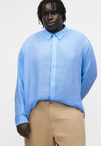 Vyras tamsia oda ir dredais, dėvintis šviesiai mėlynę su sagomis marškinius ir smėlio spalvos kelnes, stovintis priešais vienodą foną.