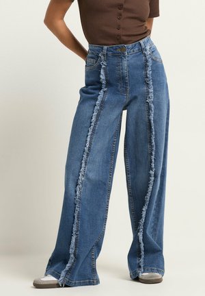 Højttaljede vidde jeans med lodrette frynsete sømme foran kombineret med en brun kortærmet top og sneakers.