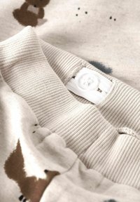 Tissu beige clair avec un col côtelé et un petit imprimé de ourson en cartoon. Comprend un bouton blanc et une texture douce.