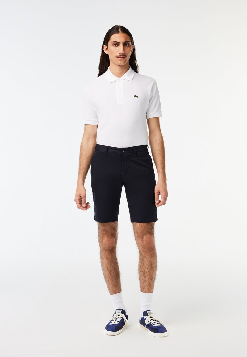 Witte polo shirt met een groen logo, gecombineerd met donkerblauwe shorts. Het model draagt witte sokken en blauwe sneakers, staande tegen een effen achtergrond.