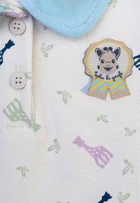 Polo in cotone con colletto azzurro chiaro, base bianca con fantasie di giraffe pastello e un badge ricamato con il testo "Sophie la girafe."